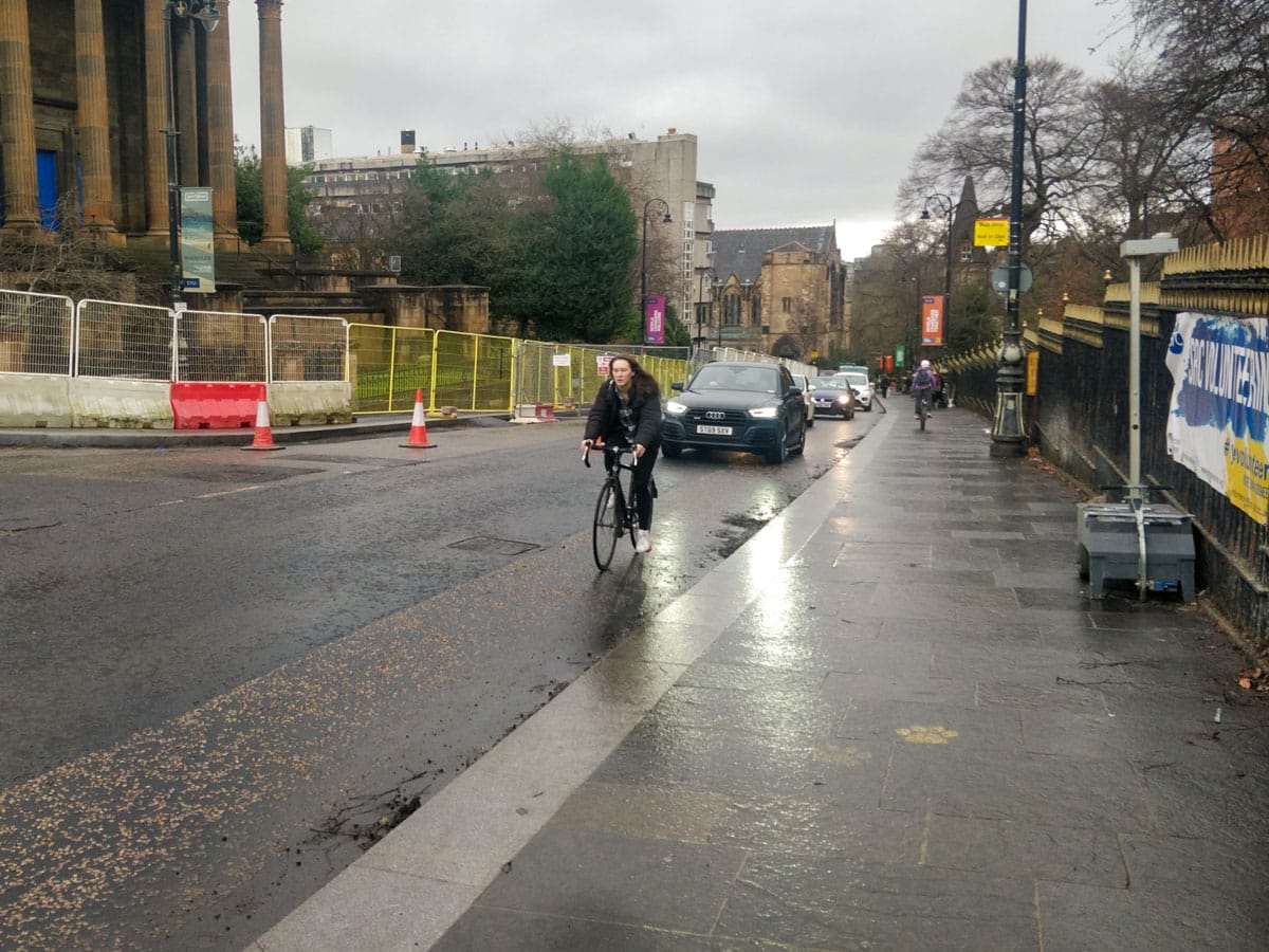 Woman cycling uphill on University Avenue with a line of cars behind (and someone cycling downhill on pavement)